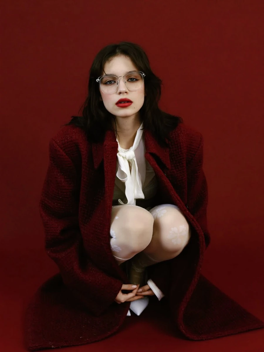 A high-fashion editorial portrait of a young woman sitting against a bold...