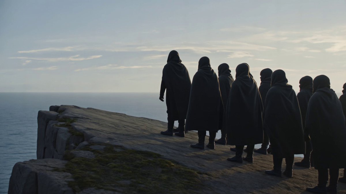 The shot opens tight on a row of cloaked figures standing along the cliff...