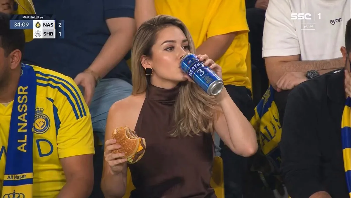 Ultra-realistic sports broadcast still of a glamorous woman sitting in a...