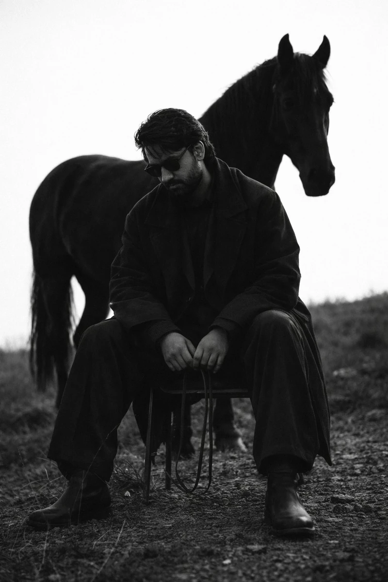 A bearded man with dark wavy hair wearing dark sunglasses, dressed in a l...