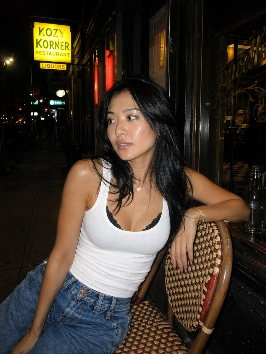 A candid nighttime street portrait of a young woman sitting casually on a...